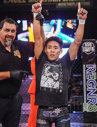 A mixed martial artist stands in a cage with both arms raised in victory while a referee holds his wrist. The athlete wears black shorts and a black t-shirt with a photo print. The background shows sponsor logos and a lit scoreboard. A mixed martial artist stands in a cage with both arms raised in victory while a referee holds his wrist. The athlete wears black shorts and a black t-shirt with a photo print. The background shows sponsor logos and a lit scoreboard.