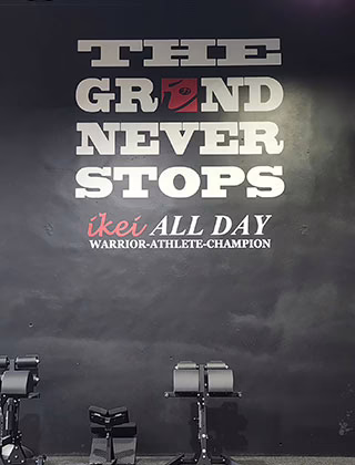 A gym wall with the motivational quote “THE GRIND NEVER STOPS ikei ALL DAY WARRIOR-ATHLETE-CHAMPION” in bold white and red letters, with workout equipment visible at the bottom. A gym wall with the motivational quote “THE GRIND NEVER STOPS ikei ALL DAY WARRIOR-ATHLETE-CHAMPION” in bold white and red letters, with workout equipment visible at the bottom.